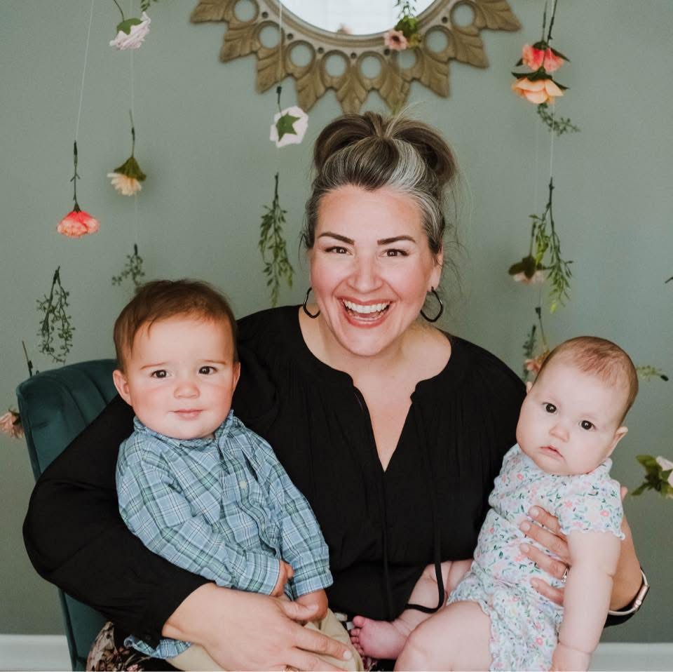 A smiling woman holds two babies, one in each arm, against a festive backdrop.