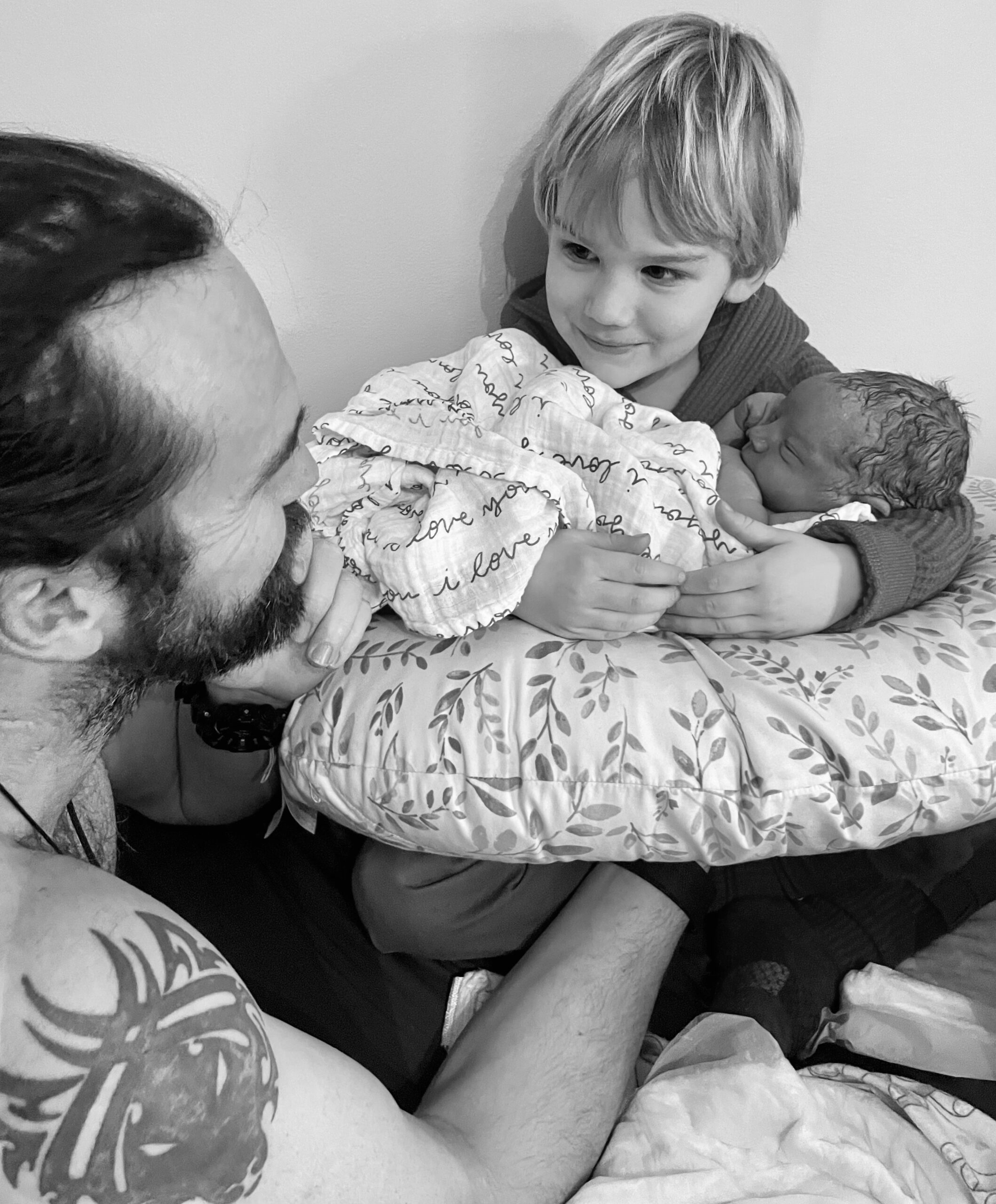 A father lovingly holds his newborn while the older sibling looks on.